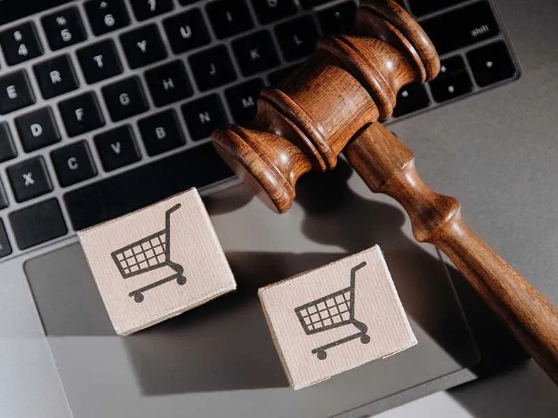 Martelo de juiz sobre o teclado de um notebook, ao lado de duas caixinhas com o ícone de carrinho de compras