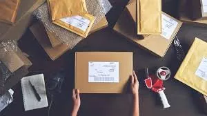 Mãos segurando uma caixa de encomenda com etiqueta, sobre uma mesa cheia de pacotes e envelopes