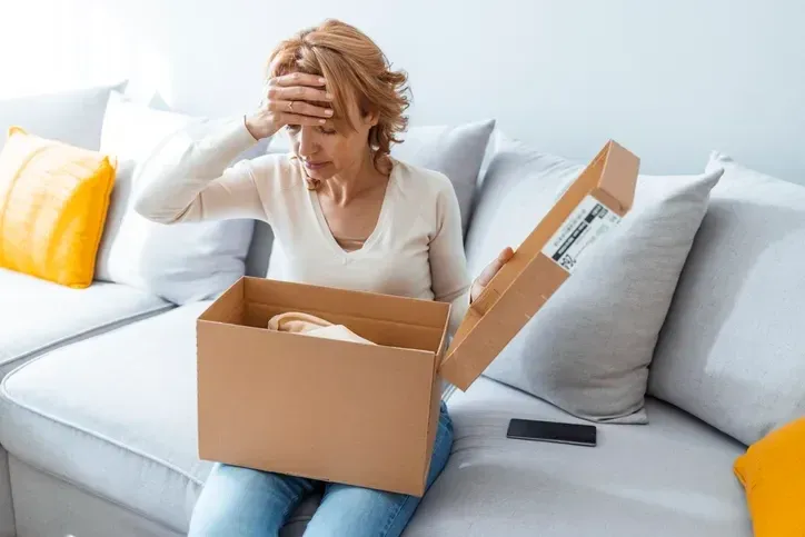 Mulher sentada no sofá, com a mão na testa, olha uma caixa de encomenda aberta no colo