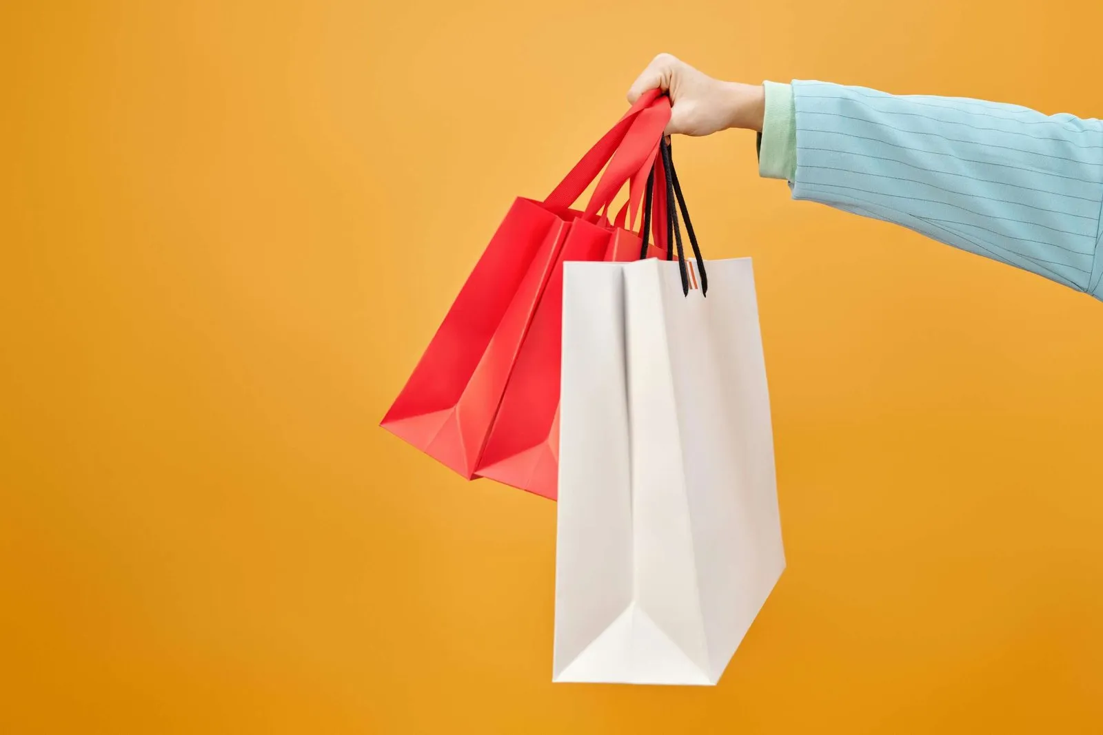Mão segurando sacolas de compras vermelha e branca sobre fundo amarelo